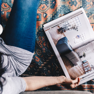 Person holding a cup of coffee while sitting on a patterned mohair rug, reading a home decor magazine. Blog discusses why mohair rugs are the latest trend in home design for their blend of luxury and sustainability.