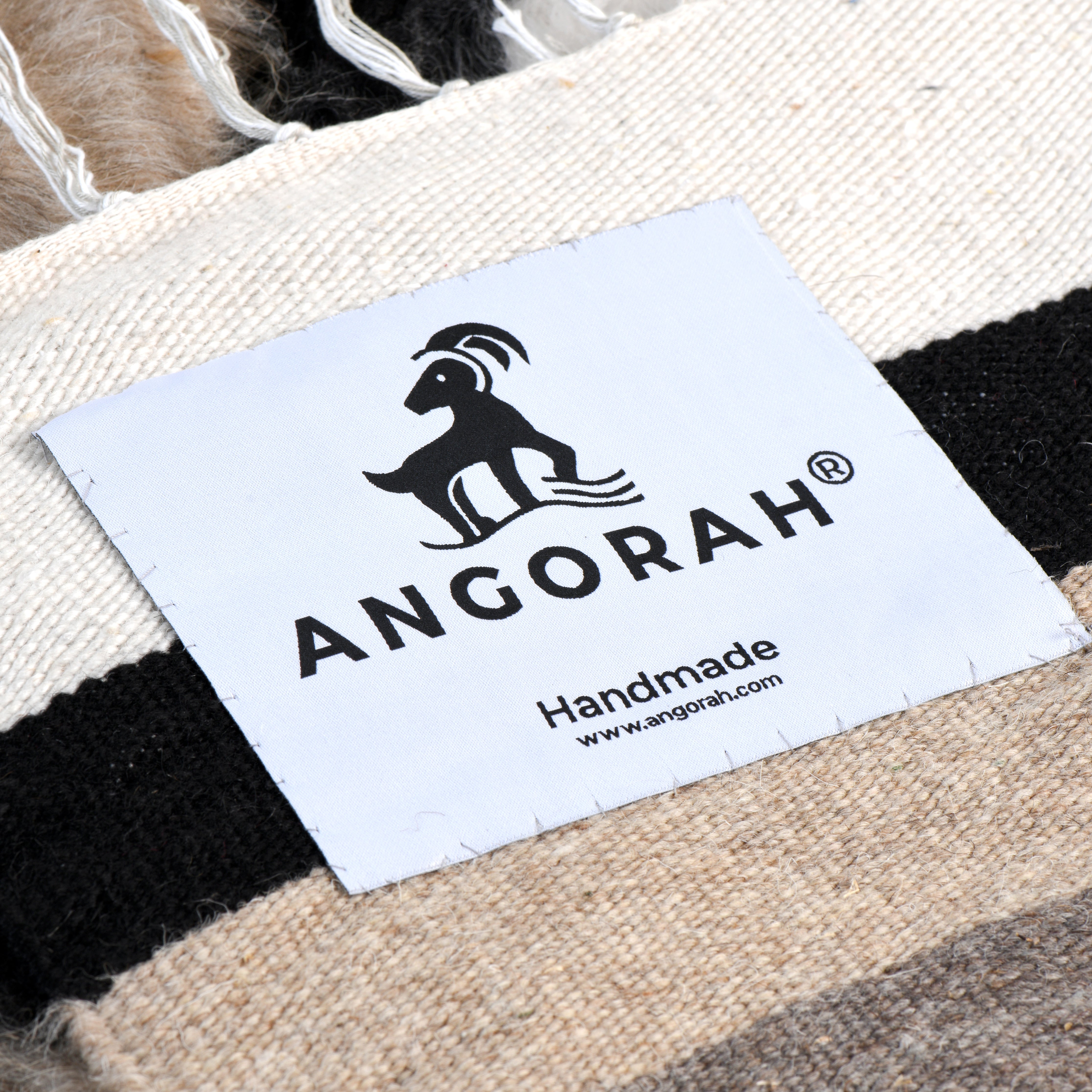 Close-up of the Angorah brand label on the GaiaPhos handcrafted rug, made from 100% organic natural mohair. The label is placed on a section of the rug featuring earthy brown, black, and white stripes, highlighting the rug's handmade quality and luxurious craftsmanship.